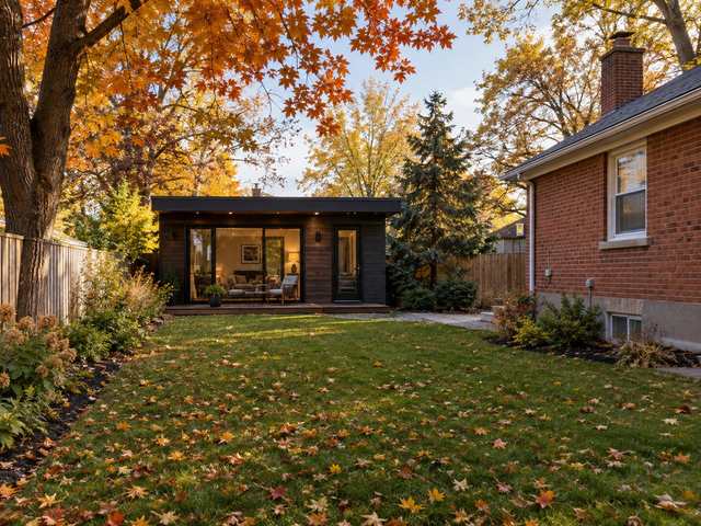 Small modern garden suite at the back of a deep lot behind a red-brick bungalow, with autumn trees on the lawn